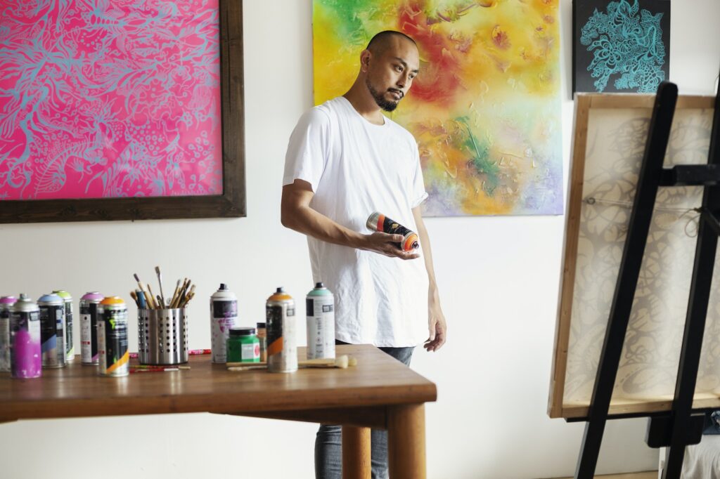 Japanese man standing in art gallery, holding can of spray paint, looking at artwork on easel.